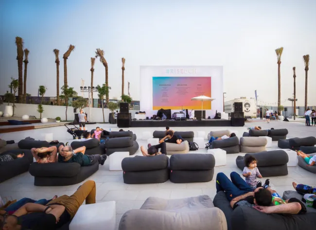 Outdoor lounge seating faces a large festival screen and stage beneath palm trees