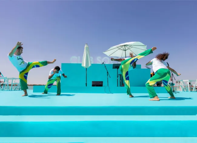 Capoeira performers leap across a turquoise outdoor stage by the beach