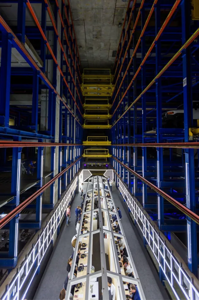 Long hospitality dinner table beneath towering warehouse vehicle racks
